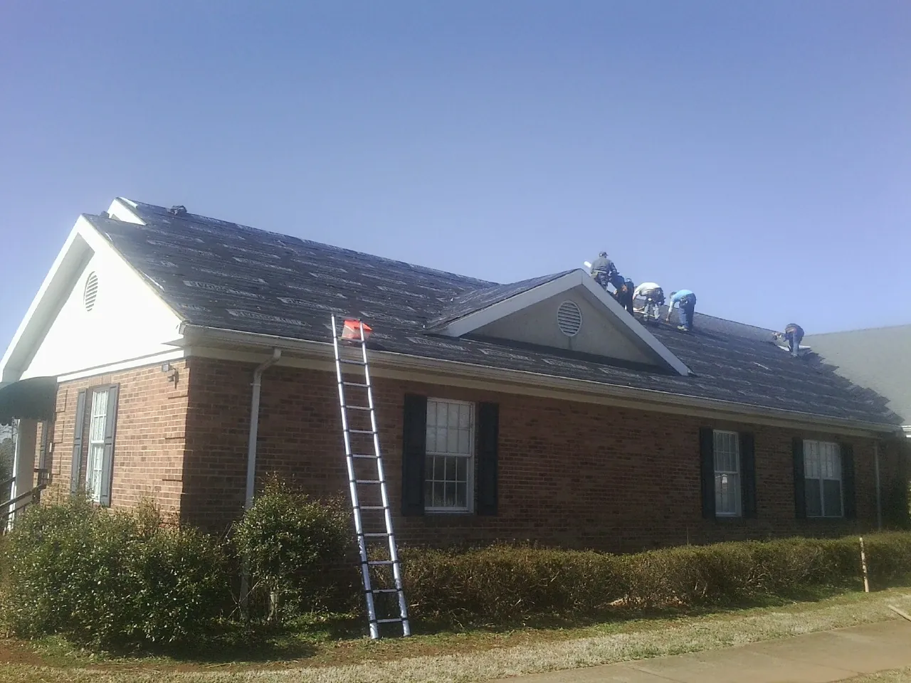 Crew tearing shingles off a brick ranch roof with a ladder set in West Sayville