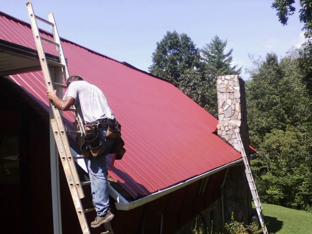 Roofer on a ladder beside a red metal roof and stone chimney — Soffit & Fascia Repair by Roofing Brothers in West Sayville